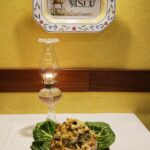 Bacalhau na broa sobre folhas de couve, com candeeiro a petróleo e o prato de faiança Cortiço Viseu