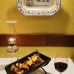 Cabrito assado com batatas em travessa de barro, copo de vinho tinto e candeeiro a petróleo, com o prato de faiança Cortiço Viseu na parede amarela