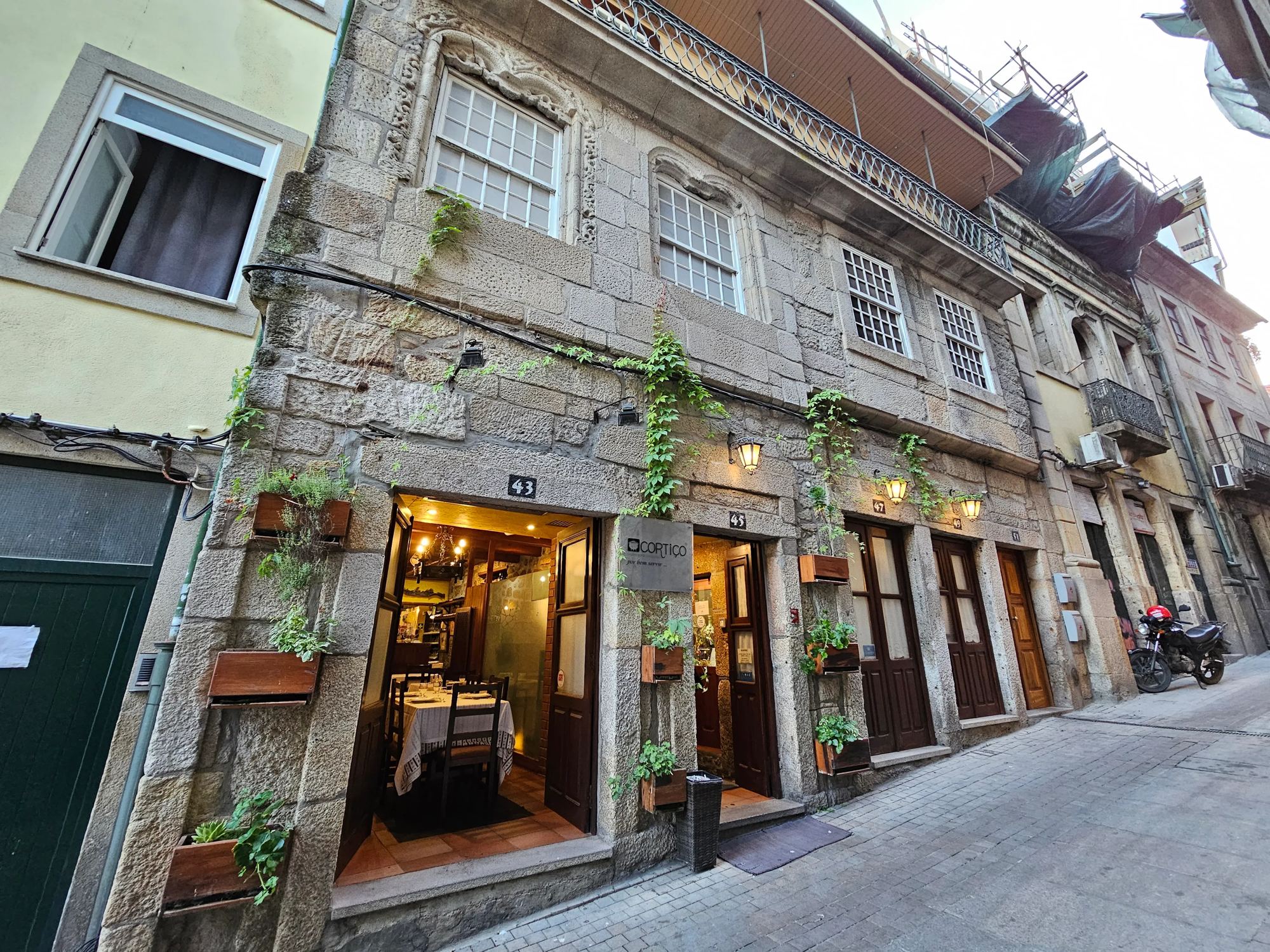 Fachada do Cortiço ao fim da tarde, com plantas na parede de granito e a porta aberta para a sala